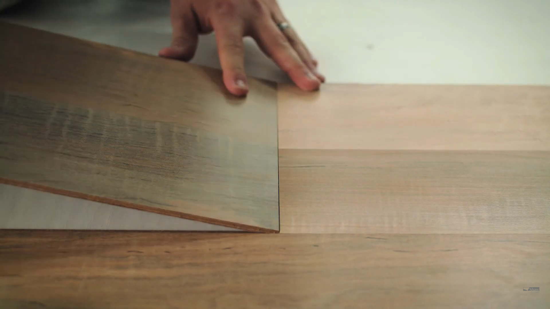 Close-up of a person installing Shaw laminate flooring by angling the tongue side of one plank into the groove of another during the floating floor installation process, showcasing the click-lock mechanism on wood-look laminate planks.