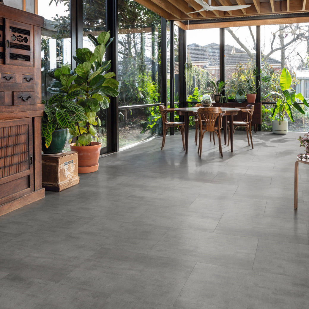 Bright indoor space with gray tiled floor, wooden furniture, and lush plants; large windows provide a view of greenery outside.
