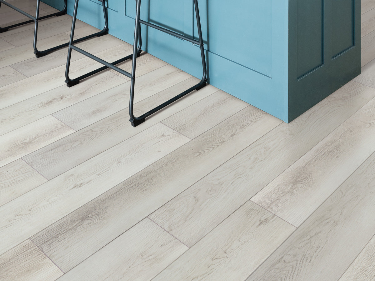 Narrow, dark board outlines lead around a blue kitchen island and black metal stools, showing Shaw Paladin Plus Feather Grey (0278V-01178) in a modern interior.