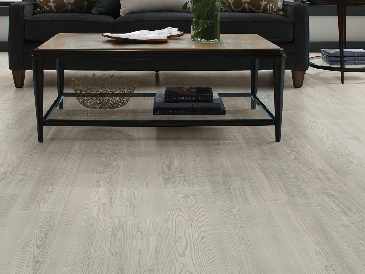 Under a wood-top coffee table with a black metal frame, Shaw Anvil Plus 20 Mil Clean Pine (2357V-05077) forms a light gray-beige floor in a modern living room with a dark sofa. Find Clean Pine's room view at Floor City for your flooring review.