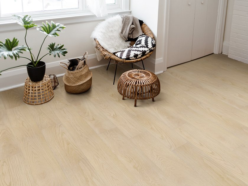 Shaw Fresh Take Lawson Khaki wood-look vinyl planks show pale beige coloring and fine grain in a window-side reading nook with a woven chair, wicker footstool and baskets.