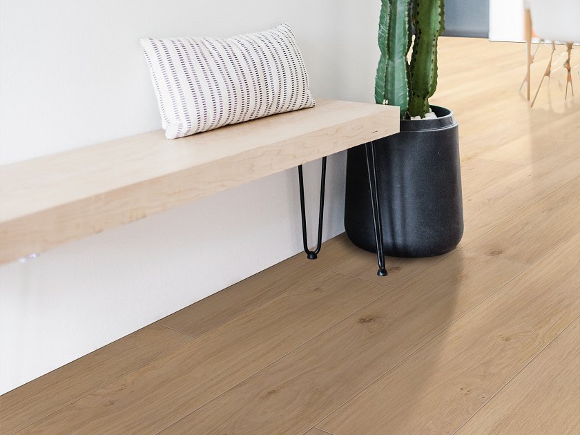 Shaw Fresh Take Occasional Beige wood-look vinyl flooring shows warm beige tones, flowing grain and small knots beside a wooden bench with a striped cushion and a tall potted cactus.