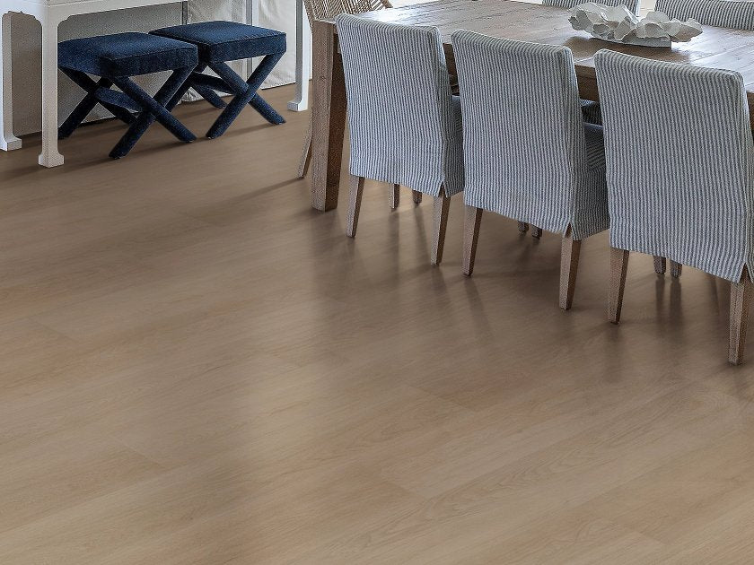 Shaw Fresh Take Bentwood Tan wood-look vinyl flooring shows muted tan-brown boards and gentle grain variation beneath a dining table with striped upholstered chairs and cross-leg stools.