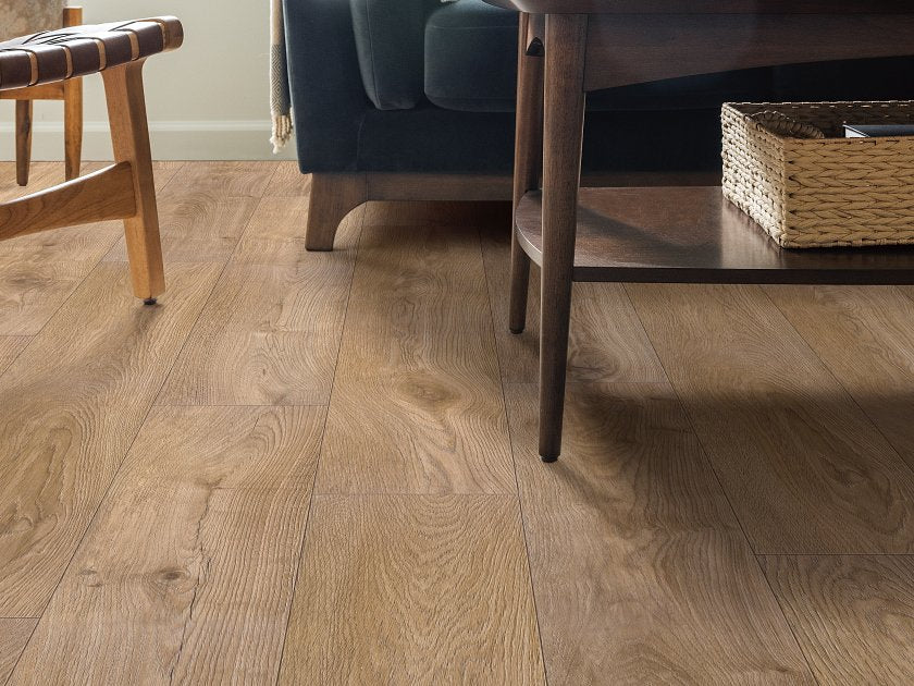 Shaw Fresh Take Cabriole Brown wood-look vinyl planks show warm brown tones, broad grain arches and dark knots beside a woven-seat chair and a dark wooden shelf table.