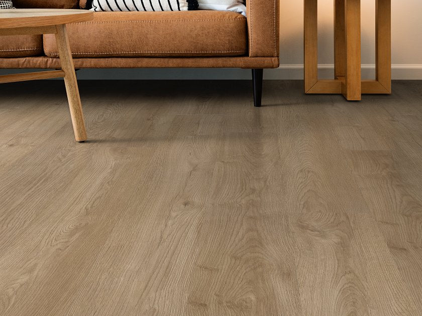 Shaw Fresh Take Wingback Brown wood-look vinyl flooring shows muted brown boards with broad grain curves and elongated knot shapes beneath a sofa beside angular wooden tables.