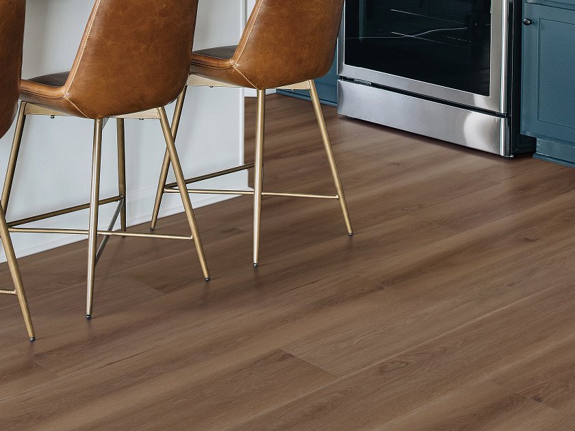 Shaw Fresh Take Ladderback Brown wood-look vinyl flooring has medium-to-dark brown boards and long grain streaks beneath upholstered kitchen bar stools beside a stainless steel range.