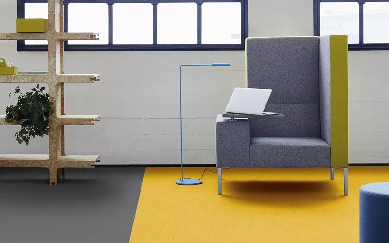 Tarkett Flooring LinoFloor modern gray and green chair on top of bright yellow flooring, there is a wooden shelving unit to the left and a round blue ottoman to the right