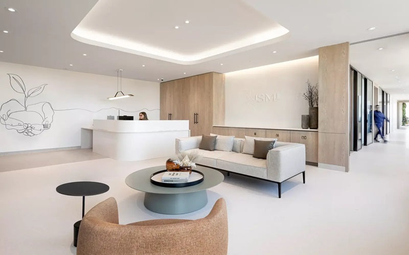 Tarkett Flooring Melodia Collection A bright and clean lobby with white flooring, walls, and ceiling. A couch, table, and chairs sit in the waiting room while a receptionist sits at a white desk in the background.