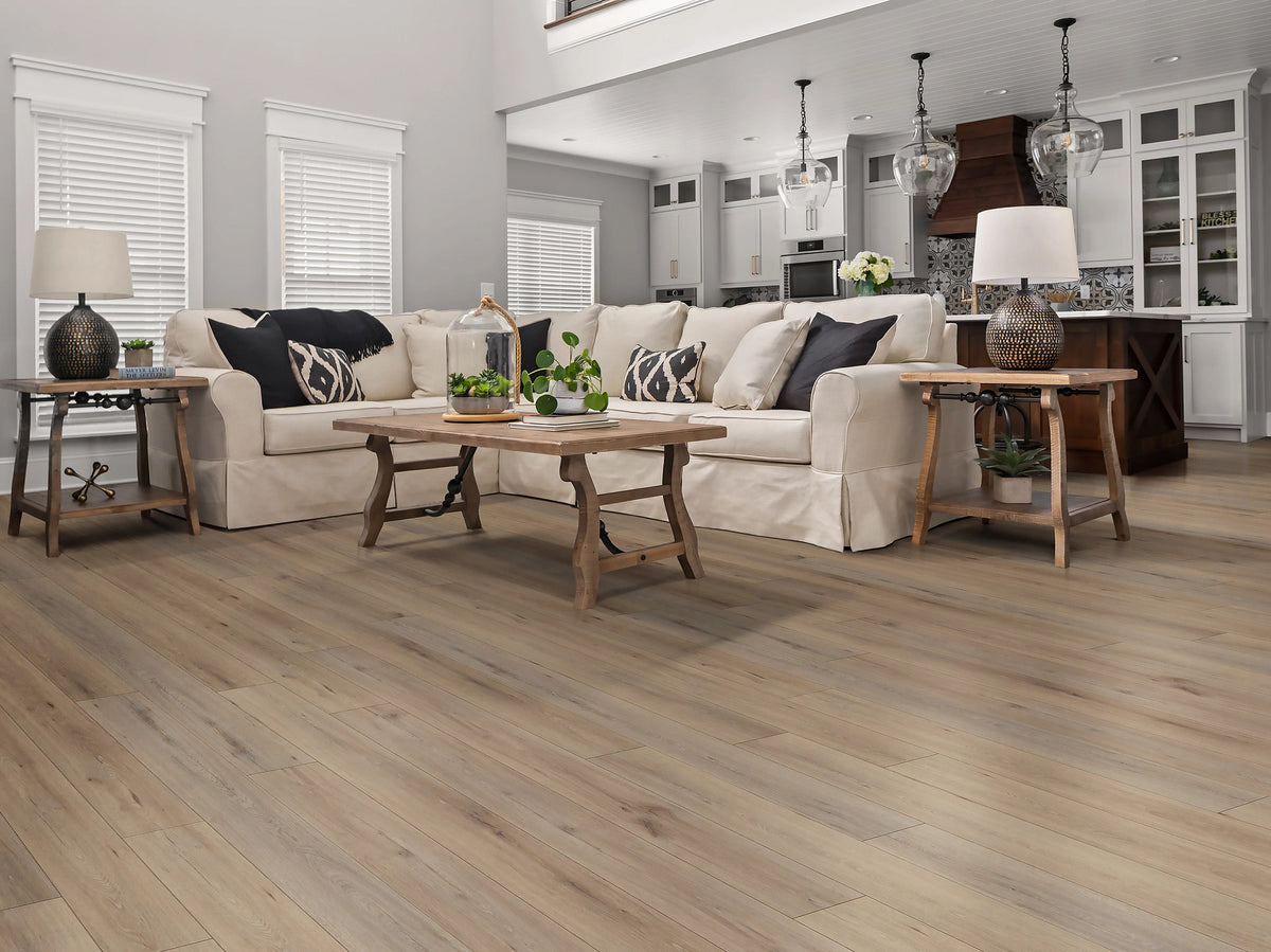 Rustic grain details extend beneath a cream sectional and wood tables in the open-plan farmhouse-style room showing Shaw Paladin Plus Wild Dunes (0278V-07089), with a kitchen beyond.