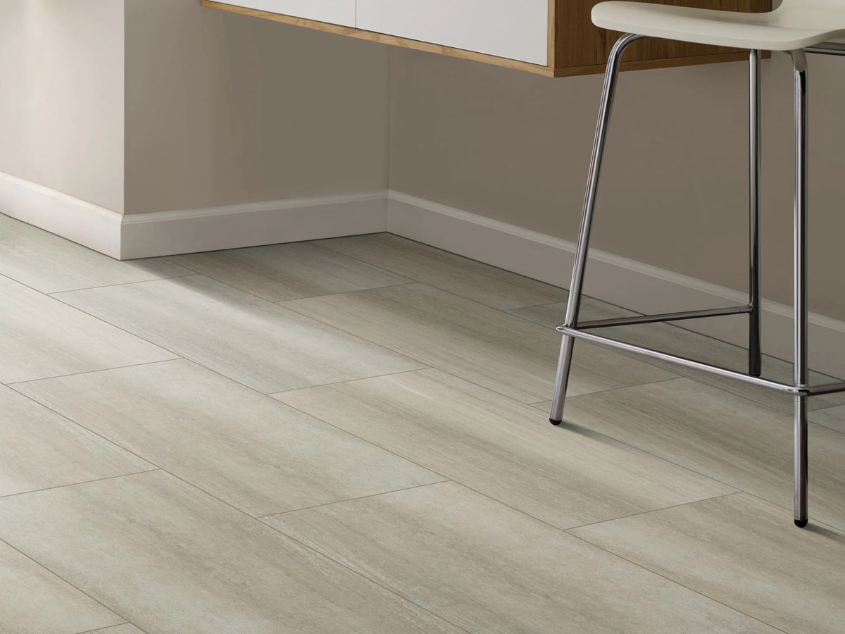A white counter stool with slender metal legs stands over Ash 1022V-01008 from Shaw Paragon Tile Plus, against taupe walls in a minimalist kitchen corner.