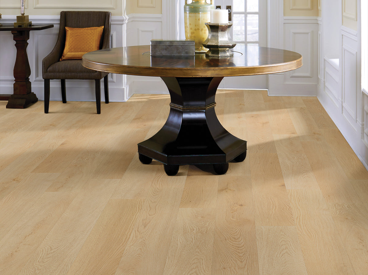 Warm honey coloring spreads around the round pedestal table in this traditional room, showing Shaw Anvil Plus 20 Mil River Bend Oak (2357V-00296) against paneled walls and an accent chair. Ask Floor City about River Bend Oak and its room view.