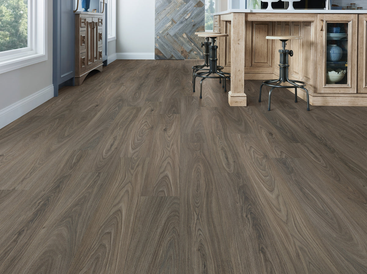 Large arched grain patterns lead across Shaw Anvil Plus 20 Mil Dark Elm (2357V-00915) toward wood bar cabinetry and metal stools, grounding the rustic room in deep gray-brown tones. Discover the room view of Dark Elm with Floor City.