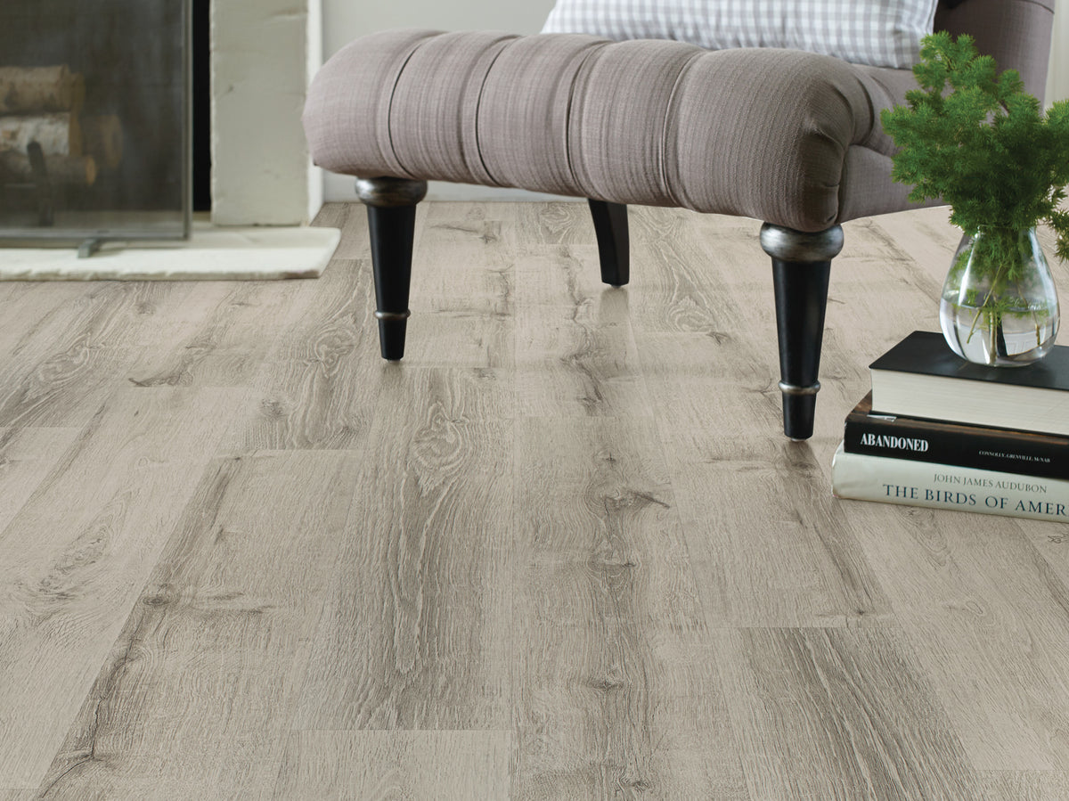 Long vinyl boards extend beneath an upholstered chair with turned legs, showing the staggered layout and gray-beige coloring of Shaw Anvil Plus 20 Mil Beach Oak (2357V-01023) in a traditional sitting area. Review Beach Oak's room view with the team at Floor City.