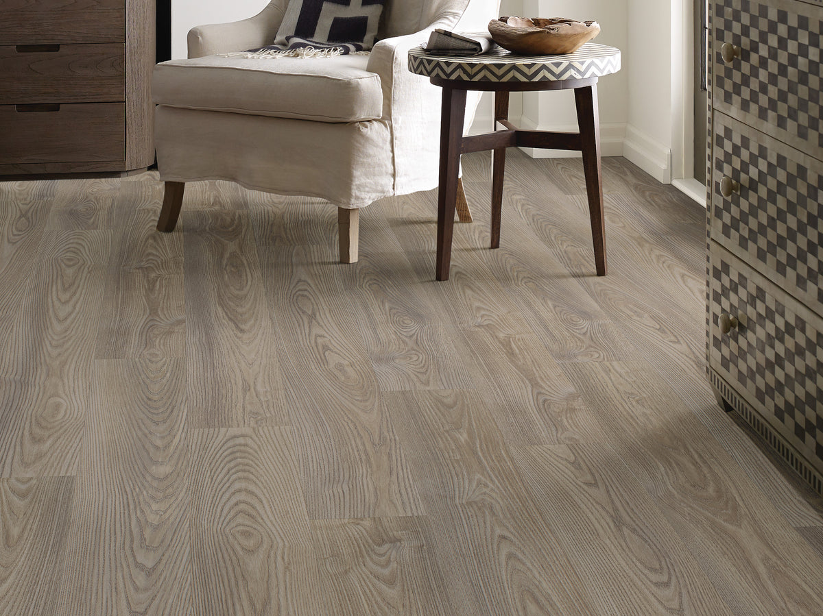 Muted gray and brown shades run beneath an upholstered armchair beside a small wooden table, showing Shaw Anvil Plus 20 Mil Grey Chestnut (2357V-07062) in a transitional reading corner. See how Grey Chestnut's room view informs your choice at Floor City.