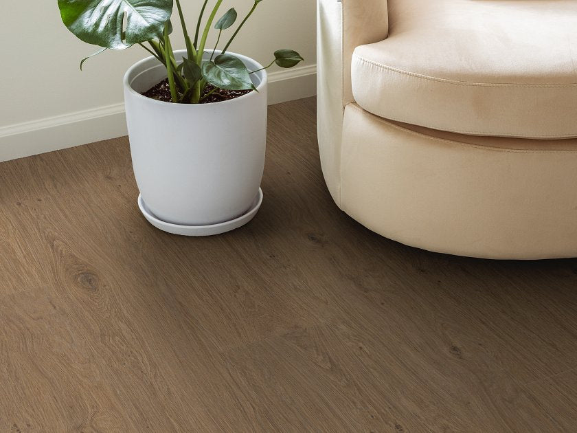Shaw Fresh Take Tuxedo Bronze wood-look vinyl planks show muted smoky brown coloring, fine grain and small knot markings beside a curved upholstered chair and a large potted plant.