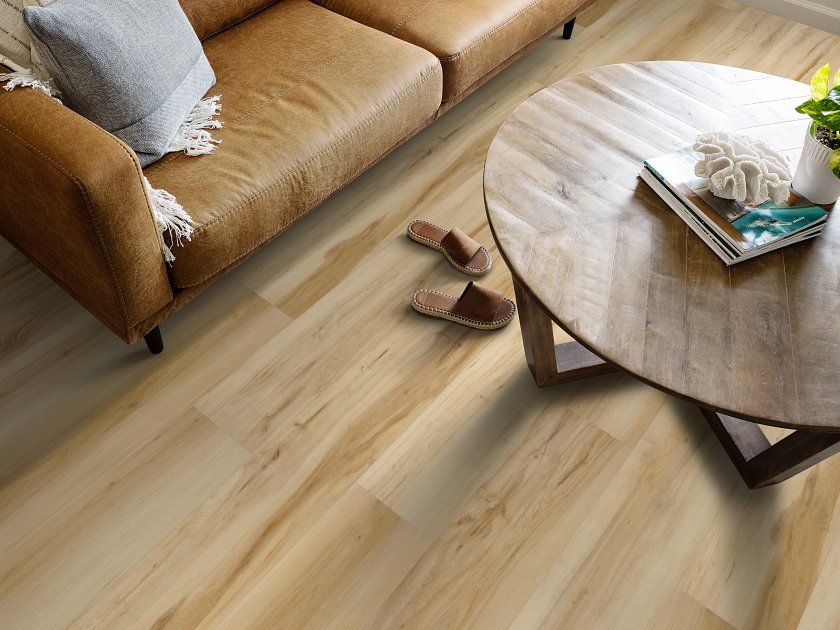 Shaw Fresh Take Butterfly White wood-look vinyl flooring shows pale cream and tan boards with darker flowing grain beneath a leather sofa and an oval wooden coffee table.