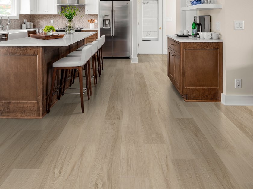 Shaw Fresh Take Lounge Greige wood-look vinyl flooring displays varied gray-beige boards and long grain streaks in a kitchen with a wooden island, upholstered stools and matching cabinetry.