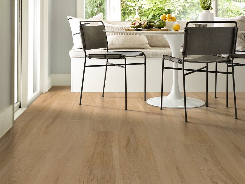 Shaw Fresh Take Chaise Tan wood-look vinyl planks show golden tan coloring and darker knot patterns beneath a round pedestal dining table with dark-framed chairs beside tall windows.
